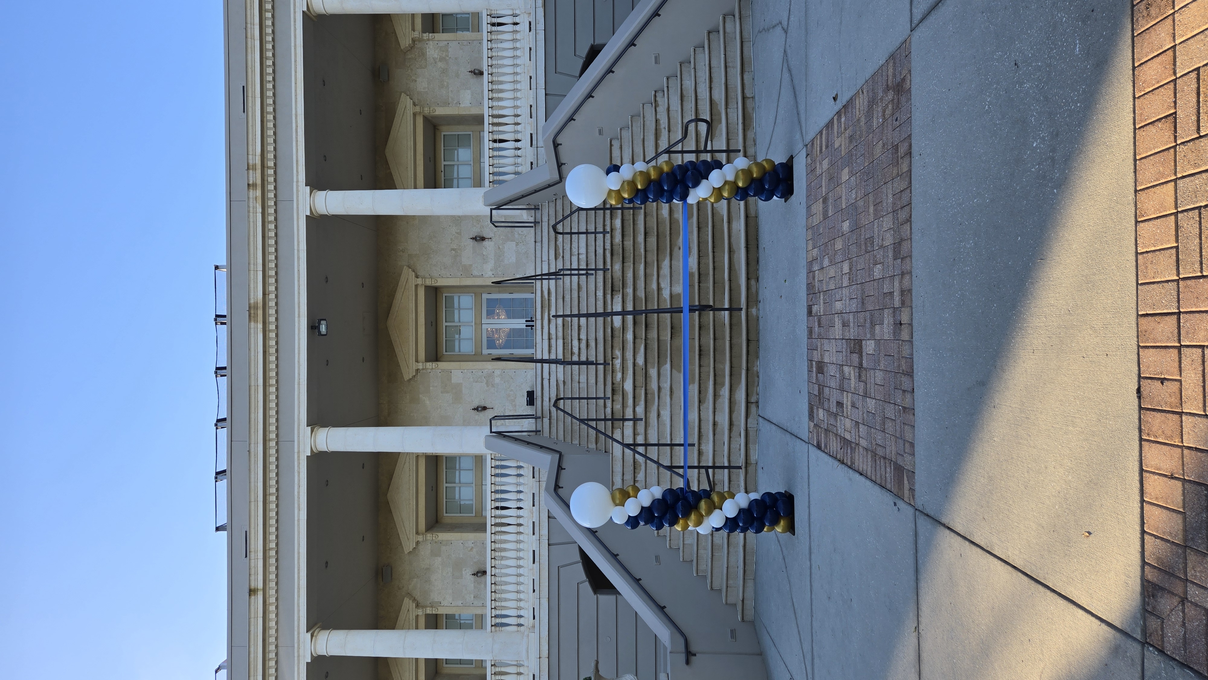 Wide view of a ribbon cutting ceremony display with two navy, gold and white balloon columns at a classic building entrance