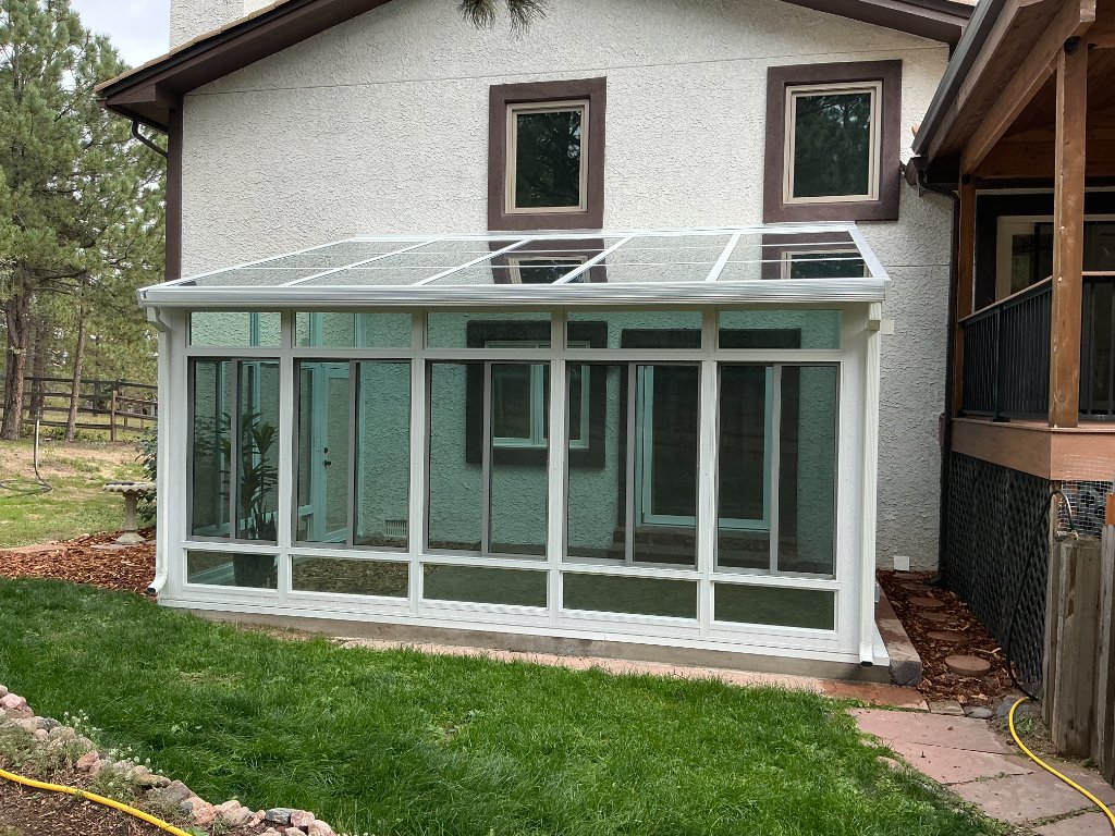 Glass solarium sunroom with a sloped shingled roof attached to a stucco home beside an elevated wooden deck