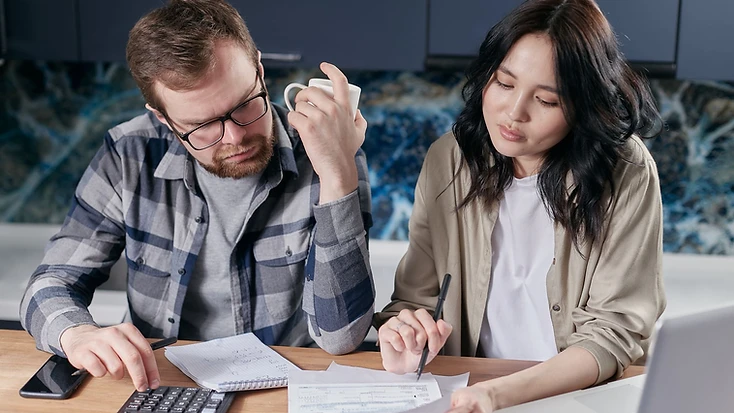 A couple reviewing household income and expenses at their kitchen table with a laptop, calculator and notebook.