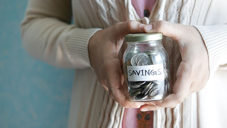 A person holding a glass jar labelled Savings, filled with coins set aside at home.