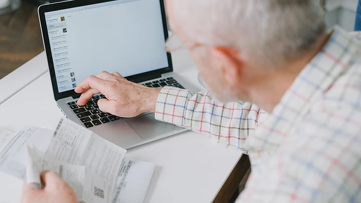 An older man using a laptop at a desk while reviewing paper receipts and financial documents.