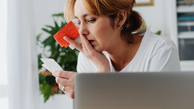 A woman at a home desk reviewing credit card statements and receipts, with a calculator and notebook beside her laptop.