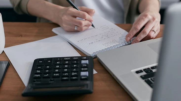 Hands writing a household budget in a notebook beside a calculator and laptop on a wooden desk.