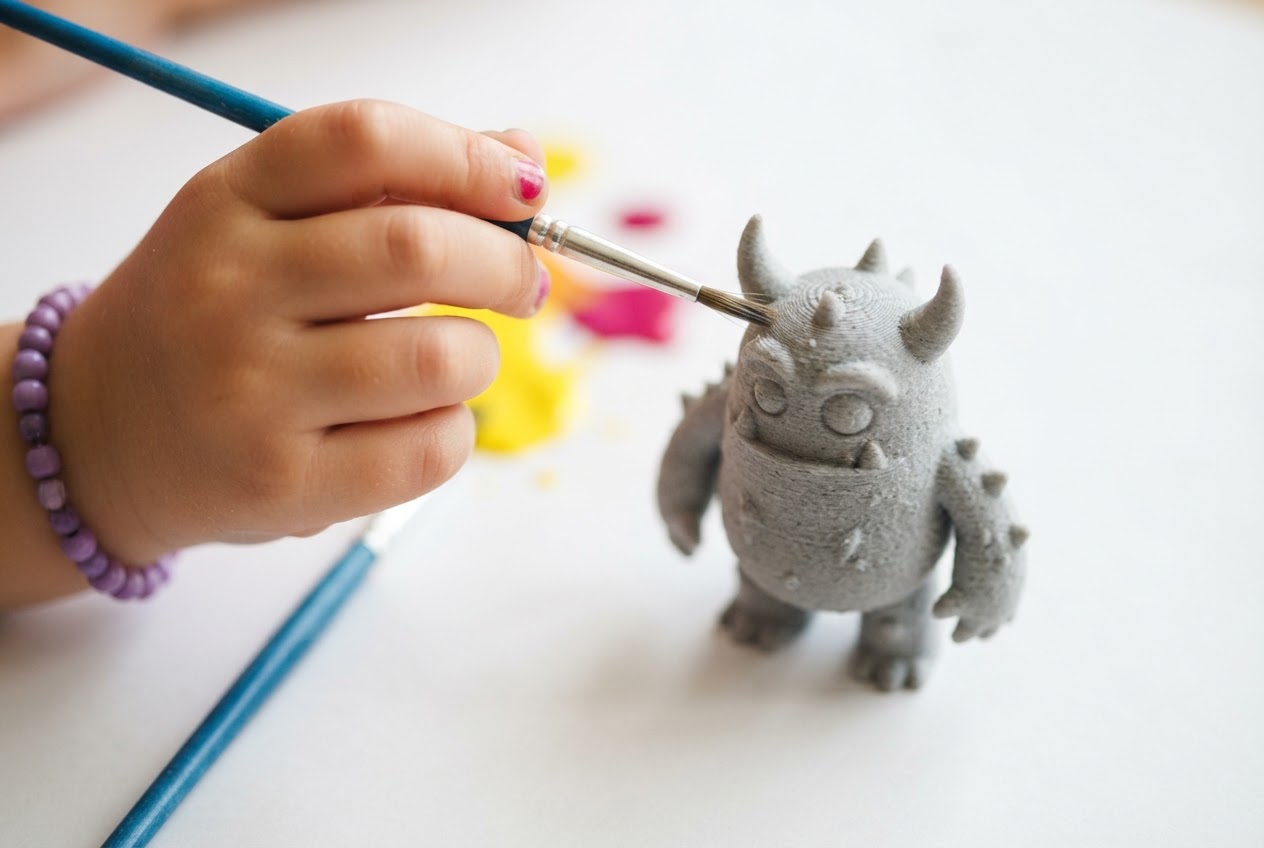A hand holding a paintbrush near a custom 3D printed grey PLA monster figurine ready to paint
