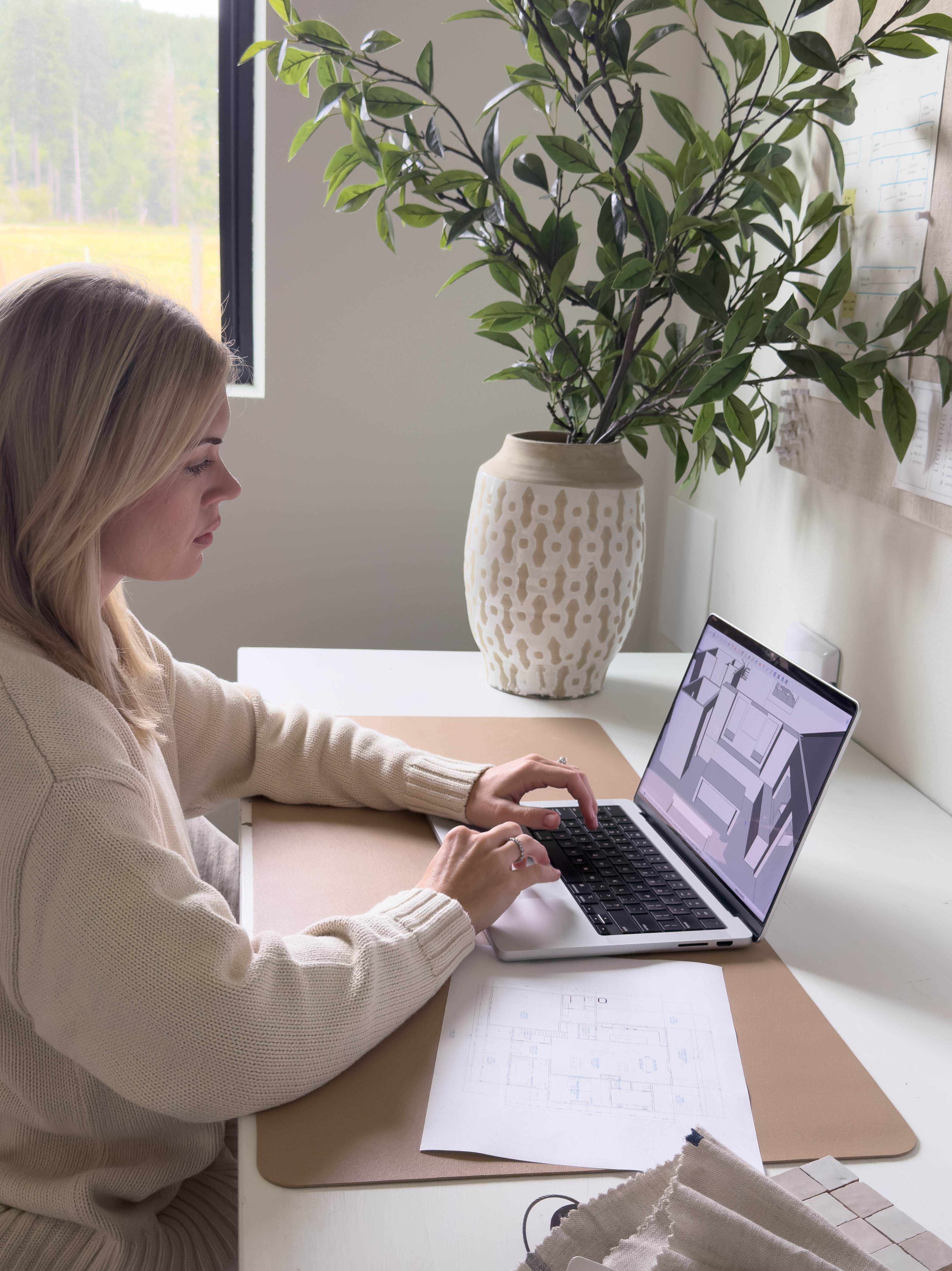 Interior designer reviewing fabric swatches and a floor plan at a wooden desk in warm window light