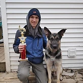 Trainer kneeling with a German Shepherd and a trophy earned at a working dog competition.
