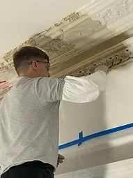 A craftsman in protective gloves and glasses stripping paint from an ornate plaster cornice, raw beige plaster emerging beneath