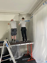Two craftsmen on mobile scaffolding taping protective plastic sheeting beneath the ornate cornice line