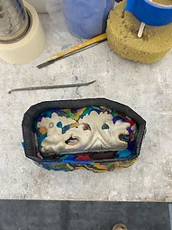 A mould-making station on the workbench, with a plaster casting bedded in coloured modelling clay inside a black mould case