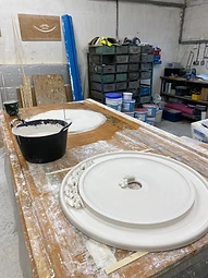 A circular plaster ceiling rose resting on a workbench beside a bucket of plaster mix and workshop shelving