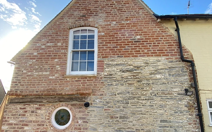 A gabled elevation in red brick and weathered stone with lime mortar joints, a sash window, and a black downpipe against a clear sky