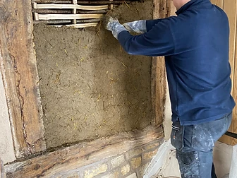 A craftsman pressing straw-bound daub by hand into a timber frame panel between aged oak beams