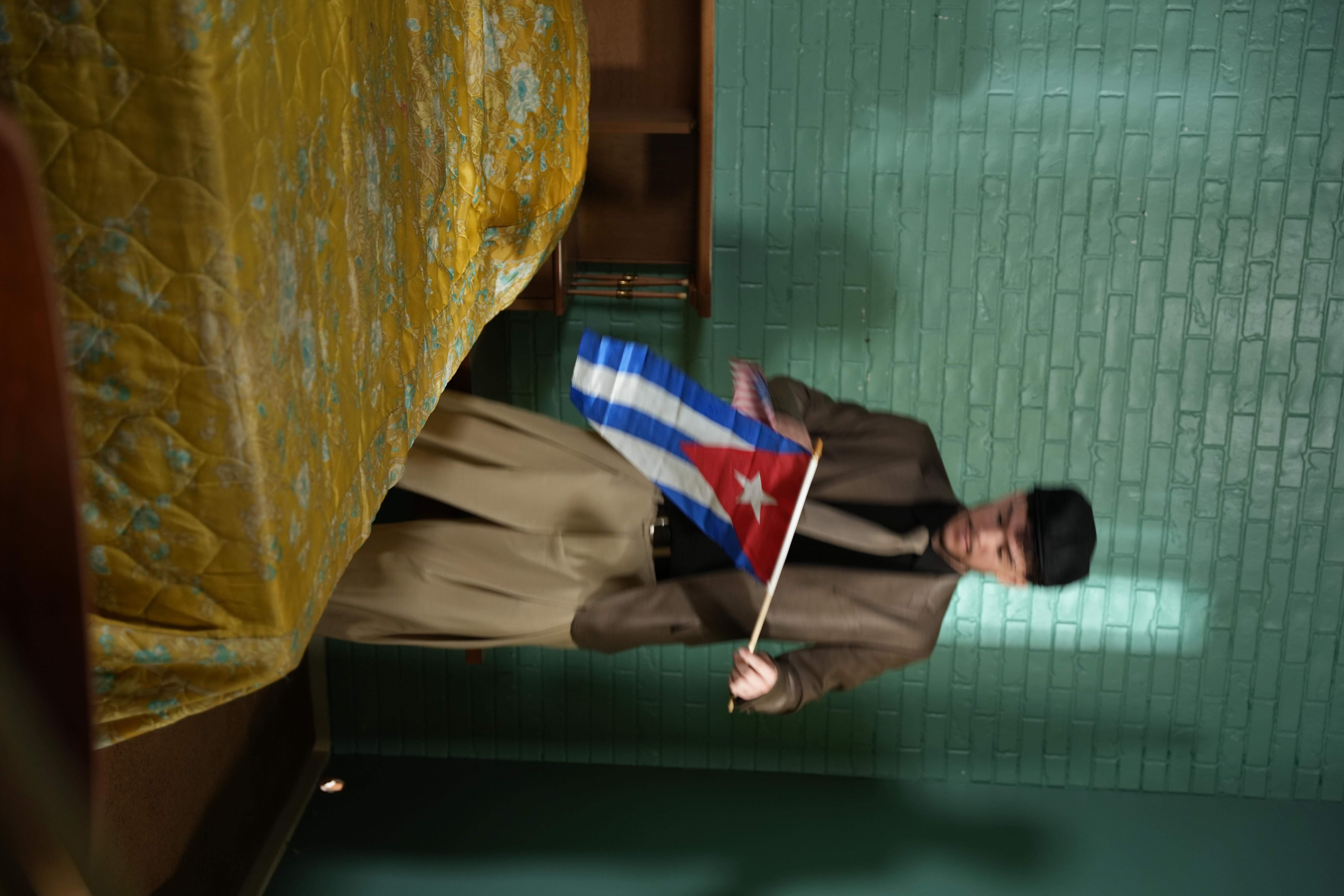 Yuma sosteniendo una bandera cubana frente a un muro de ladrillo verde