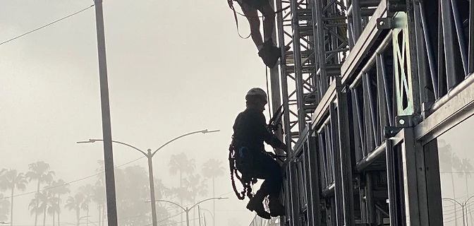 Three riggers in climbing harnesses ascending a steel scaffold truss against a foggy sky
