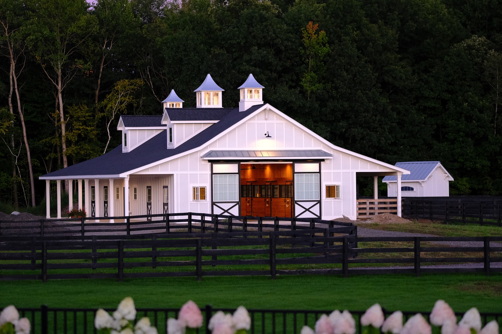 White equestrian barn at dusk with three cupolas on the roofline and warm interior light