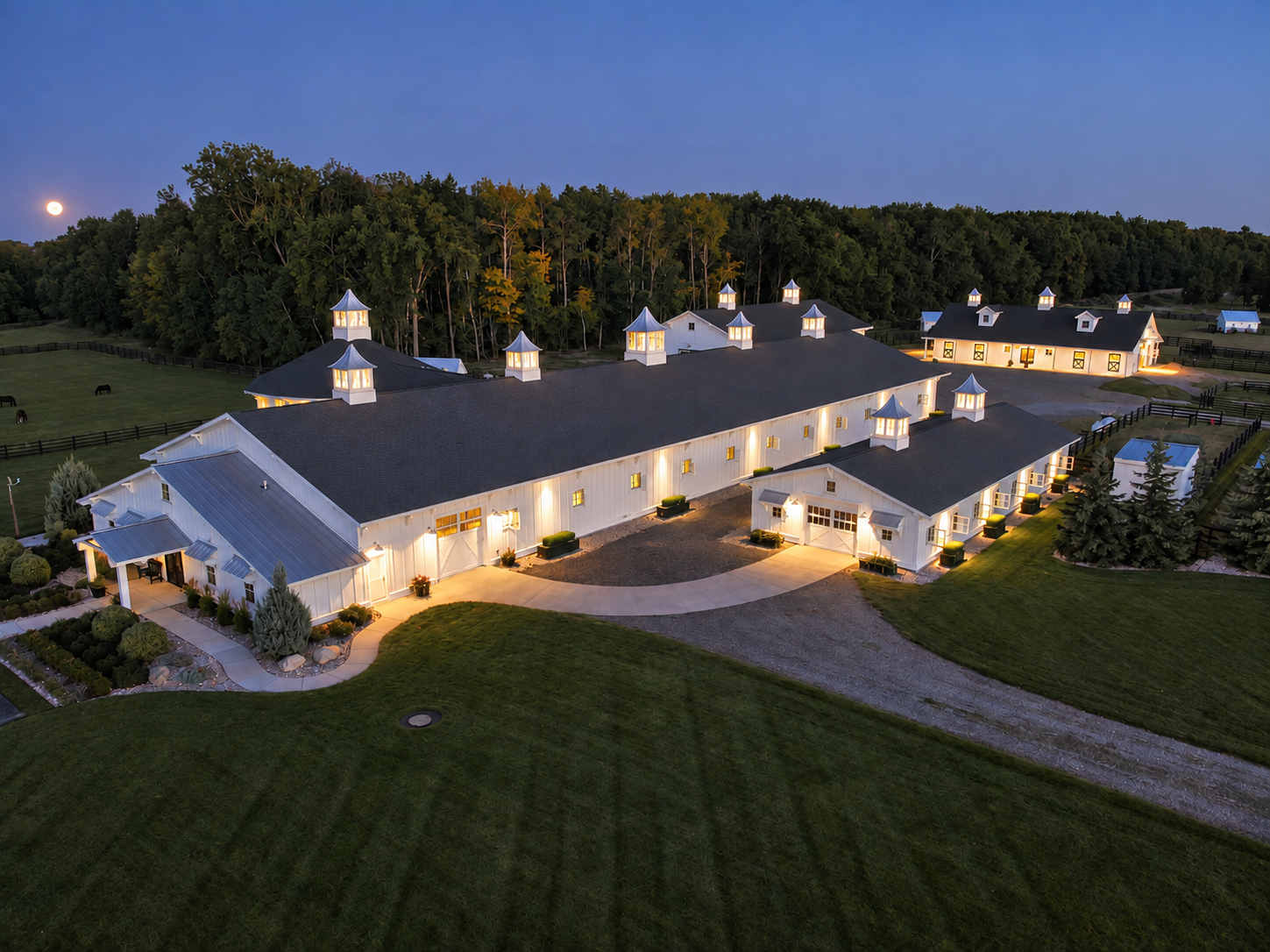 Aerial dusk view of a luxury equestrian horse barn with illuminated custom cupolas against a twilight sky