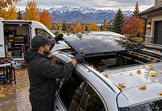 Mobile technician fitting replacement panoramic sunroof glass on an SUV in a Utah driveway, with autumn leaves and the Wasatch mountains behind
