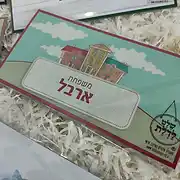שלט לדלת הבית בעיצוב אישי ומקסים של סטודיו שלט לדלת. השלט המעוצב מתאפיין בקו איורי וצבעוני, המציג מבנה בית משפחתי ציורי על רקע תכלת ושמיים עם עננים. במרכז השלט מופיע הכיתוב 'משפחת ארבל' בפונט ברור ומרשים, בתוך מסגרת בהירה ומעוטרת. השלט מתאים במיוחד כשלט לכניסה לבית, המעניק מגע אישי וחם לכל כניסה. מדובר בפריט עיצוב שלטים איכותי, שניתן להזמין כשלט אישי במגוון סגנונות. סטודיו שלט לדלת מתמחה בייצור שלטים לבית ולחדרי ילדים, המשלבים יצירתיות ואיכות, והשלט הזה הוא דוגמה מצוינת לשלט מעוצב שמוסיף חן ואופי לכל חלל.