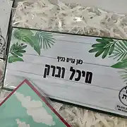 שלט לדלת הבית מעוצב ואישי מבית סטודיו שלט לדלת, המציג דוגמה לשלט אישי עם הכיתוב 'כאן גרים בכיף מיכל וברק'. השלט בעל רקע דמוי עץ בגוון לבן-אפרפר עם איורי עלים טרופיים ירוקים בפינות, המעניקים לו מראה רענן וטבעי. זהו שלט מעוצב בסגנון מודרני המתאים לכניסה לבית ומעניק מגע חם ומזמין. ניתן להזמין שלטים אישיים דומים בעיצוב שלטים מותאם אישית. השלט מוצג בתוך אריזת מתנה עם שבבי נייר, מה שמדגיש את איכות המוצר כמתנה מושלמת לזוגות או למשפחות.