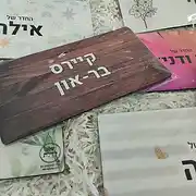 צילום תקריב של מגוון שלטים מעוצבים מבית סטודיו 'שלט לדלת', המונחים על מצע של שבבי נייר לבנים. השלטים מציגים מגוון סגנונות עיצוב של שלטים אישיים, הכוללים שלט לדלת הבית או שלט לחדר ילדים בסגנונות שונים. בולט במרכז שלט אקרילי או דמוי עץ בעל טקסטורה של עץ כהה עם הכיתוב 'קיירס בר-און' באותיות לבנות בולטות, המדגים עיצוב שלטים מודרני ואלגנטי. מסביבו שלטים נוספים: אחד עם עיטורי פרחים עדינים, שלט לחדר ילדים בגווני ורוד ותכלת עם הכיתוב 'החדר של אילה', ושלט נוסף בגווני פסים אפורים ולבנים עם לב מוזהב לכיתוב 'החדר של איה'. כל שלט מעוצב בקפידה כשלט אישי ייחודי, המתאים להתקנה כשלט לכניסה או כפריט דקורטיבי לחדרי הבית. העיצובים משלבים הדפסים איכותיים, גופנים נקיים ומראה מקצועי המעיד על רמת גימור גבוהה בעיצוב שלטים בהתאמה אישית.