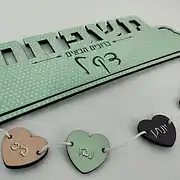 שלט עץ מעוצב ואישי לכניסה לבית, המציג את הכיתוב 'משפחת כץ' עם הכיתוב הנוסף 'ברוכים הבאים'. השלט המרכזי מגיע בצבע ירוק מנטה עדין עם דוגמת נקודות לבנות, ומחוברים אליו בחוטים לבנים מספר לבבות עץ קטנים שעליהם חרוטים שמות בני המשפחה בצבעי פסטל רכים כמו ורוד, ירוק וחום כהה. זהו שלט לדלת הבית המעניק תחושה חמימה, משפחתית ומזמינה לכל מי שנכנס. השלט מהווה דוגמה לעיצוב שלטים מקורי ומותאם אישית של סטודיו 'שלט לדלת', ומתאים במיוחד כמתנה אישית או כפריט דקורטיבי שמוסיף חן ואופי לכל כניסה לבית או לחדר.