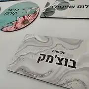 מבחר שלטים מעוצבים לבית ולמשרד מבית סטודיו 'שלט לדלת'. בתמונה מוצגים מספר שלטים אישיים בעלי סגנון עיצוב מודרני ואסתטי. במרכז בולט שלט מלבני עם טקסט 'משפחת בוצימק' על גבי רקע שיש יוקרתי בגווני אפור ולבן. בחלק העליון ניתן לראות שלט עגול עם הדפס פרחים עדין הנושא את השם 'הראל קורמן', המתאים באופן מושלם כשלט לחדר ילדים או לחדר שינה, לצד שלט נוסף עם עיצוב עלים ירוקים והשם 'אבשלום שיינפלד'. כל שלט לדלת הבית או למשרד מיוצר מחומרים איכותיים בגימור מוקפד, המעניקים פתרון עיצובי מותאם אישית לכל חלל. אם אתם מחפשים שלט אישי, שלט מעוצב בכניסה, או שלט לבית עם נגיעה של סטייל וייחודיות, הקולקציה שלנו מציעה מגוון סגנונות, החל משלט עץ ועד לשלט אקריליק, המבטיחים עיצוב שלטים ברמה הגבוהה ביותר לכל דלת.