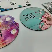 מבחר שלטים מעוצבים לבית ולמשרד מבית סטודיו שלט לדלת. בתמונה נראים שלטים בהתאמה אישית במגוון צורות וסגנונות: שלט מלבני עם הדפס פרפרים כחולים, שלט עגול בעיצוב עץ תכלת עם פרחים ורודים, ושלט עגול נוסף עם איור אמנותי של פרחי חיננית בצבעי סגול וורוד. כל שלט אישי מציג שם משפחה או טקסט ייחודי בגופן מודרני ונקי. שלטים אלו מהווים פתרון מושלם למי שמחפש שלט לדלת הבית, שלט לחדר ילדים או שלט מעוצב לכניסה. העיצובים הייחודיים מדגישים את המומחיות של הסטודיו בתחום עיצוב שלטים, ומציעים מראה אסתטי, צבעוני ומרשים שמתאים לכל חלל, בין אם מדובר בבית פרטי או כשלט למשרד, ומעניקים תחושה אישית וחמה כבר בכניסה.