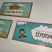 צילום מרהיב של שלושה שלטים מעוצבים לבית ולחדר הילדים מבית סטודיו שלט לדלת. השלטים מתאפיינים בעיצוב גרפי צבעוני ושמח, ומתאימים באופן מושלם כשלט לחדר ילדים או שלט לדלת הבית. השלט הימני מציג איור מקסים של ילדה מתנדנדת על ענן עם הכיתוב 'כאן גרים באהבה משפחת אמסלם' בתוך בועת דיבור לבנה. שני השלטים השמאליים מעוטרים באיור של בית חלומות צבעוני ומציגים את הכיתוב 'הבית של מאיה' בתוך תגית מעוצבת. כל שלט אישי מיוצר בעיצוב שלטים מוקפד, תוך שימוש בחומרים איכותיים שמעניקים לו מראה יוקרתי ועמיד. אם אתם מחפשים שלט מעוצב לכניסה, שלט לחדר או שלט לבית שמוסיף חמימות ואישיות לכל חלל, הדגמים הללו הם בחירה נהדרת. העיצובים משלבים גווני תכלת, אדום וניטרליים, המעניקים לשלט לדלת מראה מודרני, מזמין וייחודי.