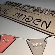 שלט עץ מעוצב לכניסה לבית בסגנון מודרני וחם. השלט המרכזי מלבני עם קצוות מעוגלים, בגוון שמנת עם הדפס נקודות עדין, ועליו הכיתוב באנגלית WELCOME ובעברית 'ברוכים הבאים' לצד המשפט 'שלום ביתנו'. תחת השלט המרכזי תלויים בעזרת חבל דקורטיבי דגלי עץ משולשים קטנים בצבעי ורוד עתיק ואפור, עליהם חרוטים שמות הילדים 'נועה' ו'תומר' באותיות לבנות. זהו שלט אישי וייחודי שמתאים לתלייה על דלת הבית או בכניסה, ומהווה דוגמה לעיצוב שלטים איכותי ומזמין לכל משפחה.