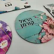 צילום תקריב של מספר שלטים מעוצבים ומותאמים אישית מסטודיו שלט לדלת. השלטים מציגים מגוון עיצובים מרהיבים, החל משלט עגול עם רקע תכלת דמוי עץ ופרחים ורודים שעליו רשום השם הראל קורמן, ועד לשלט מלבני ארוך המעוטר בפרפרים כחולים עם הכיתוב משפחת נבו. כל שלט מעוצב הוא יצירת אמנות ייחודית, המתאימה כשלט לדלת הכניסה, לחדר הילדים או כשלט לבית המעניק מראה אישי וחמים. הסטודיו מתמחה בתחום עיצוב שלטים עם דגש על איכות ועמידות, תוך שימוש בחומרים המקנים לכל שלט אישי מראה מודרני ומושקע. בין אם אתם מחפשים שלט לבית או שלט משרדי, הקולקציה המגוונת מציעה סגנונות עיצוביים שונים שמתאימים לכל חלל ומטרה.