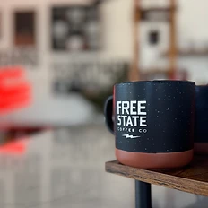 Speckled black Free State Coffee Co mug with lightning bolt logo on a wooden riser