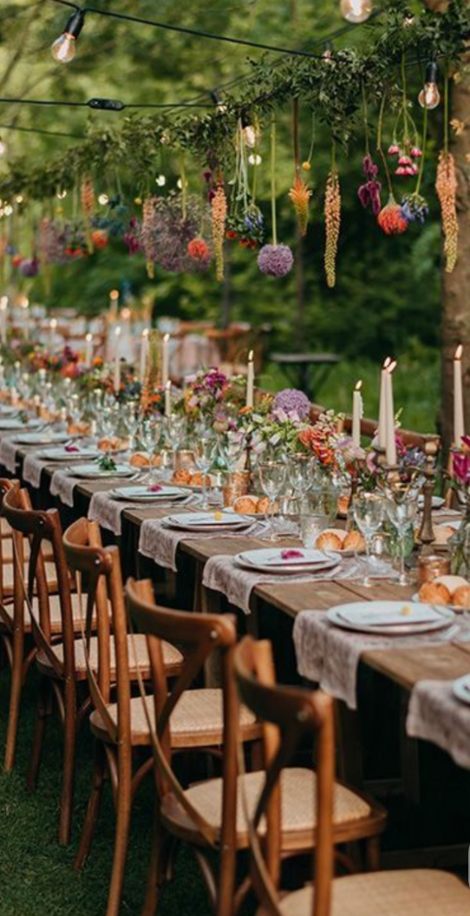 A long communal table set beneath hanging blooms and Edison light