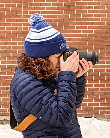 Kristel Hayes in a navy puffer and striped beanie, camera to her eye, photographing against a red brick wall in light snow