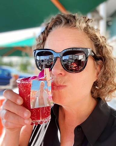 Kristel Hayes in black sunglasses holding a red drink with a Ken photo clipped to the glass, pouting playfully at the camera