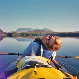 Kristel Hayes lying back in a kayak on a calm lake in 2004, a yellow dry bag at the bow, paddle across her lap, rolling hills reflected on the water