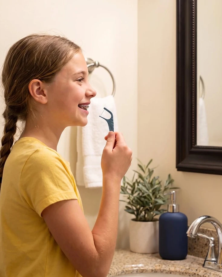 Girl with braces using a blue orthodontic floss pick in the bathroom mirror