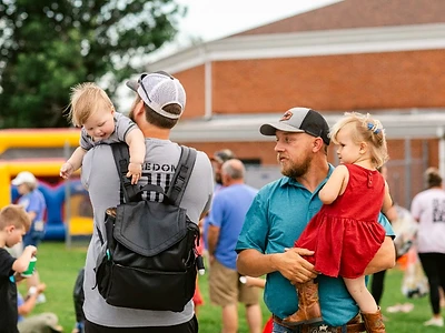 Dads hold their little ones on the lawn during an outdoor church event with a bounce house