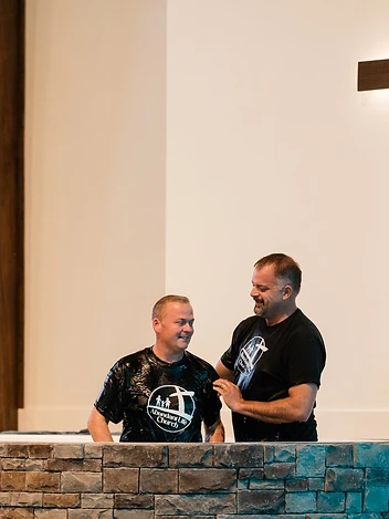 Two men stand in the baptismal pool beneath a wooden cross during a baptism celebration