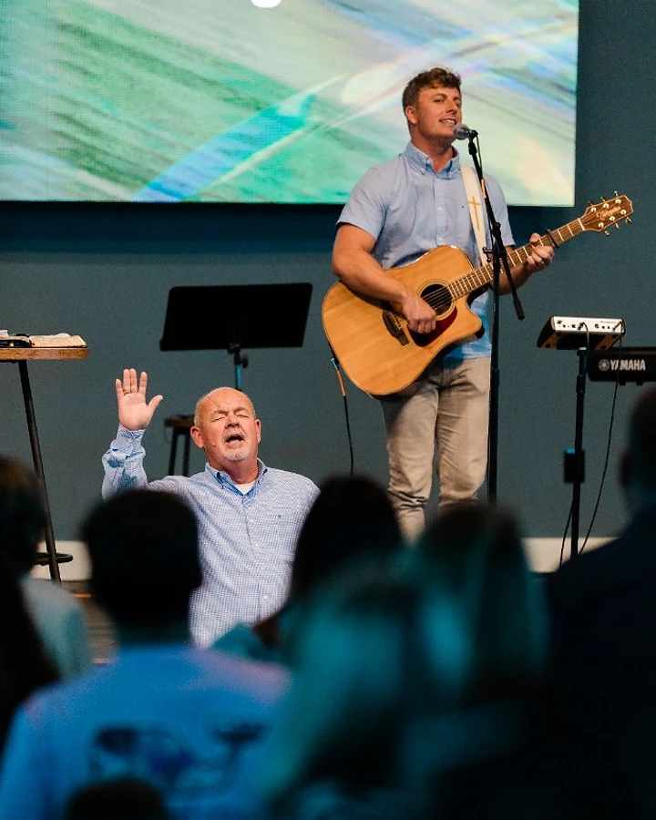 A worship leader plays guitar and sings while a man in the congregation raises his hand in praise