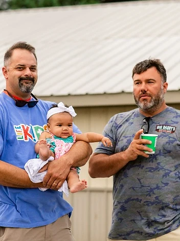 Two dads chat over coffee while one holds a baby at an outdoor gathering