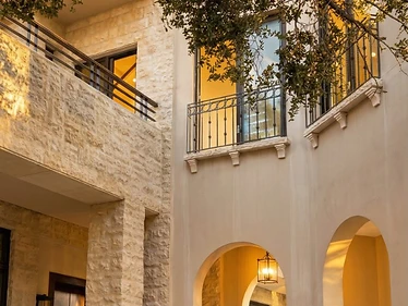 Hotel Courtyard at the Dead Sea featuring pale limestone walls, arched passages, dark timber doors, metal balconies, and a mature tree canopy