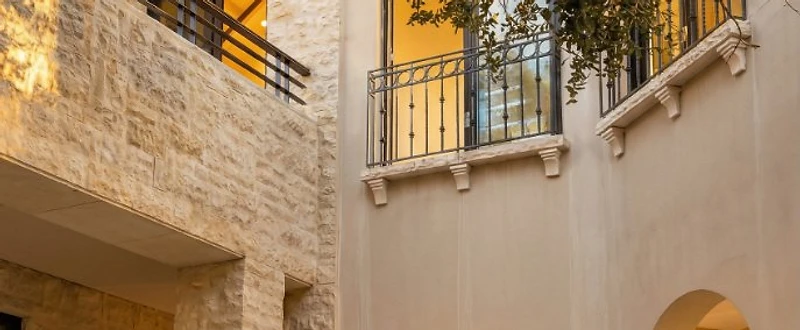 Hotel Courtyard at the Dead Sea featuring pale limestone walls, arched passages, dark timber doors, metal balconies, and a mature tree canopy