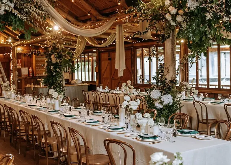 Waldara Farm wedding reception — long banquet tables with white linen and floral centerpieces inside a rustic timber-framed barn with warm string lights at Waldara Farm, Oberon