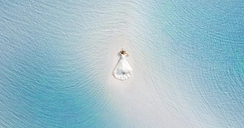 Aerial view of a bride in a white wedding dress lying on a sandbar surrounded by turquoise water