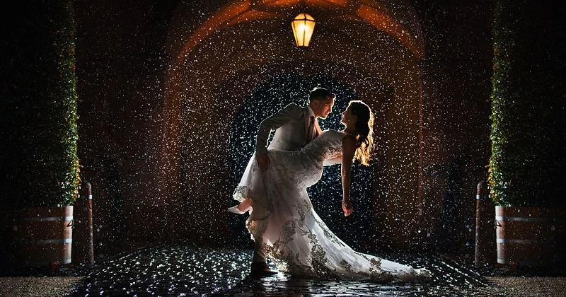 A romantic wedding couple in a dramatic dip pose under a stone archway in Sydney during a rain shower, captured with cinematic rim lighting and sparkling raindrops
