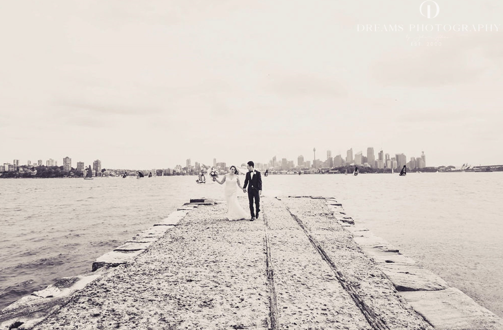 Bride and groom walking hand-in-hand along a stone jetty at Athol Hall with the Sydney city skyline in the background.