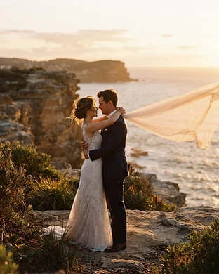 Sydney wedding photography by Dreams Photography — a couple embracing on a rocky clifftop at golden hour with a flowing veil backlit by warm sunset light