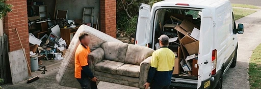 Two workers carrying an old couch and a mattress out of a garage and loading them into a removal van