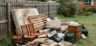 Backyard household rubbish including an old mattress, a dismantled bed frame, a worn couch and broken furniture stacked on the grass