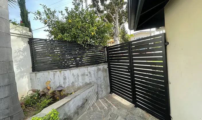 Underside of a black horizontal metal slat pergola cover over a wood-composite deck with white gravel ground and stone coping wall.